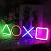Neon signs shaped like PlayStation controller buttons are displayed in a room with flowers and pineapples in the background. Produktbild-Vorschau