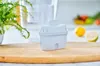 A white water filter cartridge sits on a wooden cutting board, with a blurred background of a pitcher and fruit. Náhled obrázku produktu