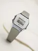 A digital watch with a silver-toned case and a gray strap is shown against a light-colored, textured background. Produktbild-Vorschau