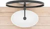 A top-down view of a white oval basin sink with a black faucet and a black circular frame above it, placed on a wooden surface. Náhled obrázku produktu