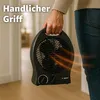 A person is holding a black electric heater with a handle in a room. Produktbild-Vorschau