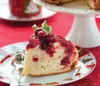 A slice of cranberry cake is presented on a festive plate with a spoon, set against a red tablecloth. A cake stand with the rest of the cake is visible in the background. Voir les images du produit