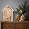 A wooden advent calendar shaped like a house is displayed on a wooden dresser, next to a vase with a Christmas tree branch and decorations. Náhled obrázku produktu