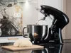 A black stand mixer with a stainless steel bowl is shown in a kitchen setting, with a ball of dough on a wooden cutting board in the foreground. Náhled obrázku produktu