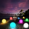 Mehrere leuchtende LED-Lichtkugeln in verschiedenen Farben auf einem sandigen Strand in der Dämmerung, mit einem Esstisch und Palmen im Hintergrund Anteprima dell’immagine prodotto