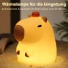 A glowing capybara-shaped night light sits on a fluffy surface, with a book and plant in the background. Produktbild-Vorschau