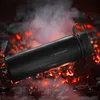 A black thermos bottle is placed on glowing embers and coals, with smoke rising around it. Produktbild-Vorschau