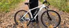 A person stands next to a white electric bicycle on a path covered with fallen leaves. Produktbild-Vorschau