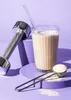 A glass of protein shake, a scoop of protein powder, and a dumbbell are arranged on purple platforms against a purple background. Produktbild-Vorschau