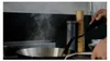 A person is cooking in a kitchen, with steam rising from a pan on a stovetop. Produktbild-Vorschau