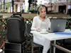 A woman is working on her laptop at an outdoor cafe table, with a backpack and suitcase next to her. Produktbild-Vorschau