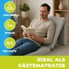 A man is reading a book while sitting on a folded mattress in a living room. Produktbild-Vorschau