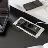 A close-up view of a black power strip with two outlets, one plugged into a black power cord and the other with a white USB cable plugged into it. The power strip is mounted in a silver frame on a black surface. A smartphone is visible in the background, c Anteprima dell’immagine prodotto