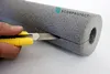 A hand is cutting a piece of grey pipe insulation with a yellow utility knife. Produktbild-Vorschau