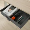 A fabric drawer organizer under a desk, containing a magazine, a pen, a pencil, and a small orange bottle. Voir les images du produit