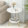 A round, two-tiered side table with a marble-like finish and gold legs, placed next to a sofa in a living room setting. It is decorated with a vase of flowers, a cup and saucer, a book, and perfume bottles. Anteprima dell’immagine prodotto