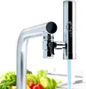 A water filter attached to a kitchen faucet, with water flowing through it and blurred vegetables in the foreground. Produktbild-Vorschau