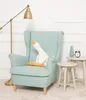 A plush goose toy sits on a light blue armchair next to a floor lamp and small side tables. Náhľad obrázku produktu