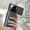 A hand holding a black smartphone case with a ring holder and a sliding camera cover. The background is a blurred interior of a room. Produktbild-Vorschau