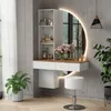 A modern vanity set with a round LED mirror, shelves, and a stool, decorated with flowers and plants. Voir les images du produit