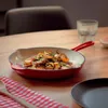 A cast iron pan filled with roasted potatoes sits on a wooden table, next to a glass of water and a red and white checkered napkin. Produktbild-Vorschau