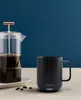 A black Ember mug and a glass French press filled with coffee are displayed on a table against a dark blue background. Náhled obrázku produktu