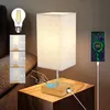 A bedside lamp with a fabric shade is shown on a wooden nightstand, with a smartphone charging on the side and an illustration of touch controls. Náhľad obrázku produktu