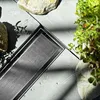 A close-up view of a modern shower drain with a brushed steel finish, surrounded by natural elements like rocks and a small plant. Anteprima dell’immagine prodotto