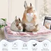 Two plush bunny toys are sitting on stacked pet blankets, with a vase of flowers on the left and a framed product information card on the right. Below the blankets are icons and text describing product features. Produktbild-Vorschau