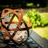 A rusty metal garden sphere sits on a dark wooden surface, with blurred green foliage in the background. Voir les images du produit