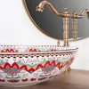 A close-up shot of a decorative ceramic sink with a brass faucet and a round mirror in the background. Náhled obrázku produktu