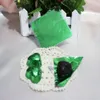 Chocolates wrapped in green foil are displayed on a white doily, with a small white picket fence and greenery in the background. Produktbild-Vorschau