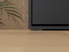 A close-up view of the base of a dark grey television or cabinet on a light wood floor, with a hint of a plant and wood-patterned wall in the background. Náhled obrázku produktu