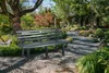 A park bench sits in a garden with a stone pathway. Produktbild-Vorschau