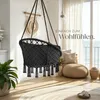 A black macrame hanging chair with tassels is shown in a bright, airy room with a window overlooking the ocean. The room is decorated with plants, cushions, and a rug, creating a cozy and relaxing atmosphere. Náhled obrázku produktu