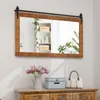 A rectangular mirror with a wooden frame and a sliding barn door hardware is mounted on a wall above a wooden dresser. The dresser holds a vase of pink tulips, a light blue storage box, and a beige handbag. Podgląd zdjęcia produktu