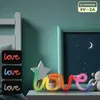 A neon LED 'LOVE' sign is displayed on a nightstand next to a bed, with a framed picture of a night sky in the background. Anteprima dell’immagine prodotto
