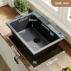 A black granite kitchen sink is shown with dimensions and features labeled. It is installed on a wooden countertop with white cabinets below. Produktbild-Vorschau