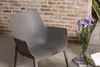 A grey chair is placed next to a small table with flowers and a book, with a brick wall in the background. Produktbild-Vorschau