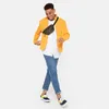 A man wearing a yellow jacket, white shirt, blue jeans, and sneakers, with a green fanny pack across his chest, posing against a white background. Produktbild-Vorschau