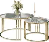 Two round coffee tables with gold colored frames and glass tops. The larger table on the left has a vase of white flowers on it. The smaller table on the right has a candle and two cups of coffee on it. Produktbild-Vorschau
