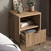 A wooden nightstand next to a bed, with a clock and plant on top. Voir les images du produit