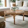 A round wooden coffee table with a glass top, featuring a small vase with eucalyptus leaves, placed in a living room setting with rattan furniture and white cushions. Podgląd zdjęcia produktu
