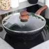 A black frying pan with a glass lid and a wooden handle sits on a black induction stovetop. Náhled obrázku produktu