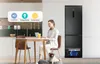 A woman is working on a laptop at a table in a modern kitchen, with a refrigerator in the background and a dog sitting at her feet. Produktbild-Vorschau