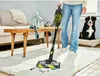 A person is vacuuming a rug in a living room with a dog sitting on its bed in the background. Produktbild-Vorschau