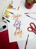 A flat lay of art supplies including pens, scissors, a brush, and a mug, with a handwritten calligraphy quote on paper. Voir les images du produit