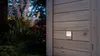 An outdoor motion sensor light is mounted on a wooden gate at dusk, with plants in the background. Produktbild-Vorschau