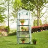 A small greenhouse with shelves filled with potted plants sits on a grassy lawn in a garden setting. Produktbild-Vorschau