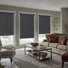 A living room scene with three windows covered by dark gray roller blinds, a sofa, a coffee table, and an armchair. Produktbild-Vorschau